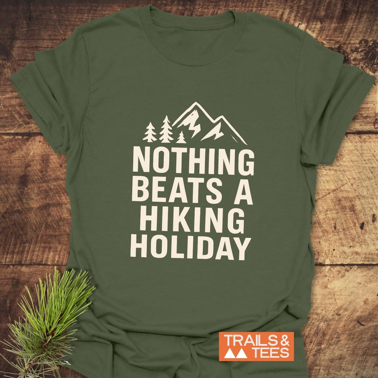 Nothing Beats A Hiking Holiday T-Shirt featuring bold white text and a mountain icon, laid flat with a pine sprig beside it for styling.
