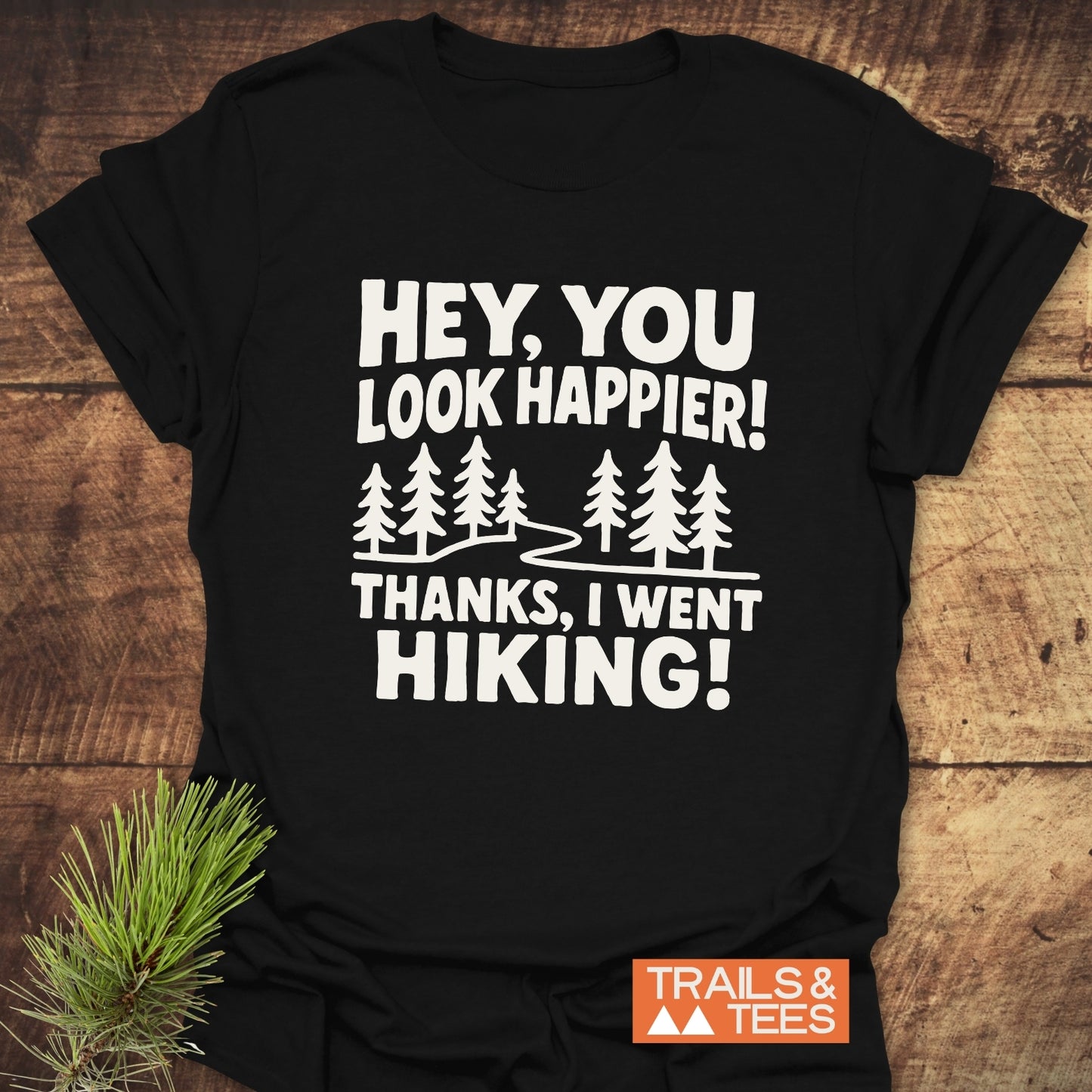 Hey, You Look Happier T-Shirt with white text, winding trail graphic, and 'TRAILS & TEES' logo, on wood surface, with pine sprig beside.