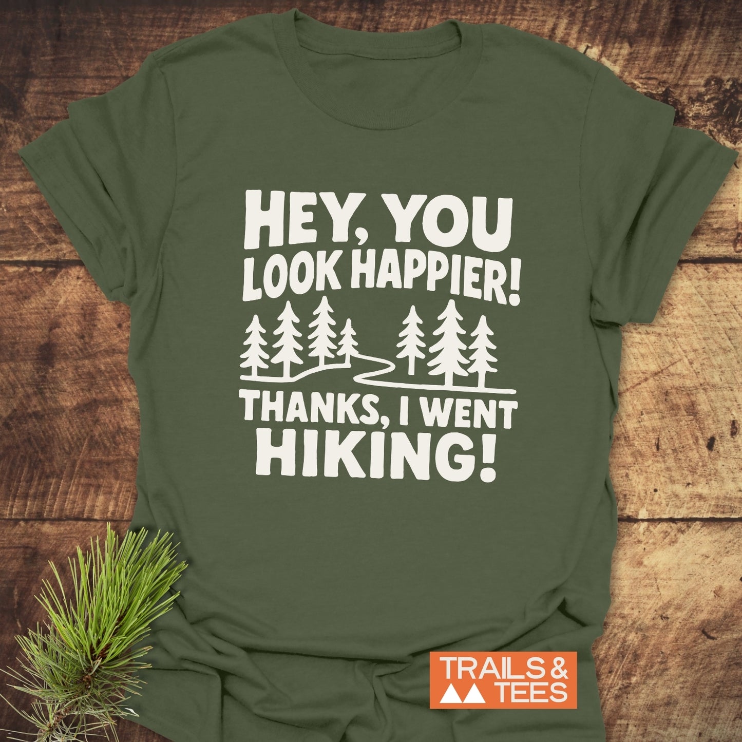 Hey, You Look Happier T-Shirt displaying bold white text and trail graphics, crafted from soft cotton, laid flat with a pine sprig beside it.