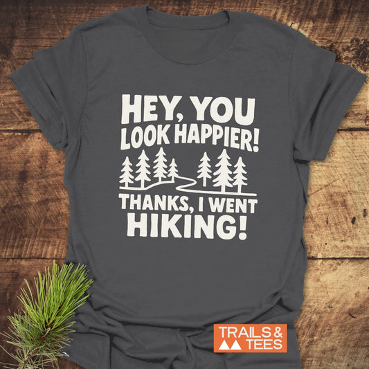 Hey, You Look Happier T-Shirt: Charcoal-gray tee with white hiking-themed graphic, pine trees, and Trails & Tees logo. Soft cotton, pre-shrunk, durable print.