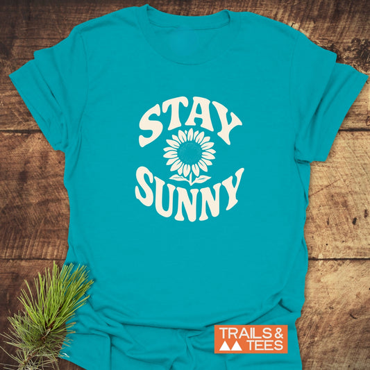 Stay Sunny T-Shirt with STAY SUNNY and sunflower graphic, showcasing soft cotton and durable print, styled with pine sprig and Trails & Tees logo.