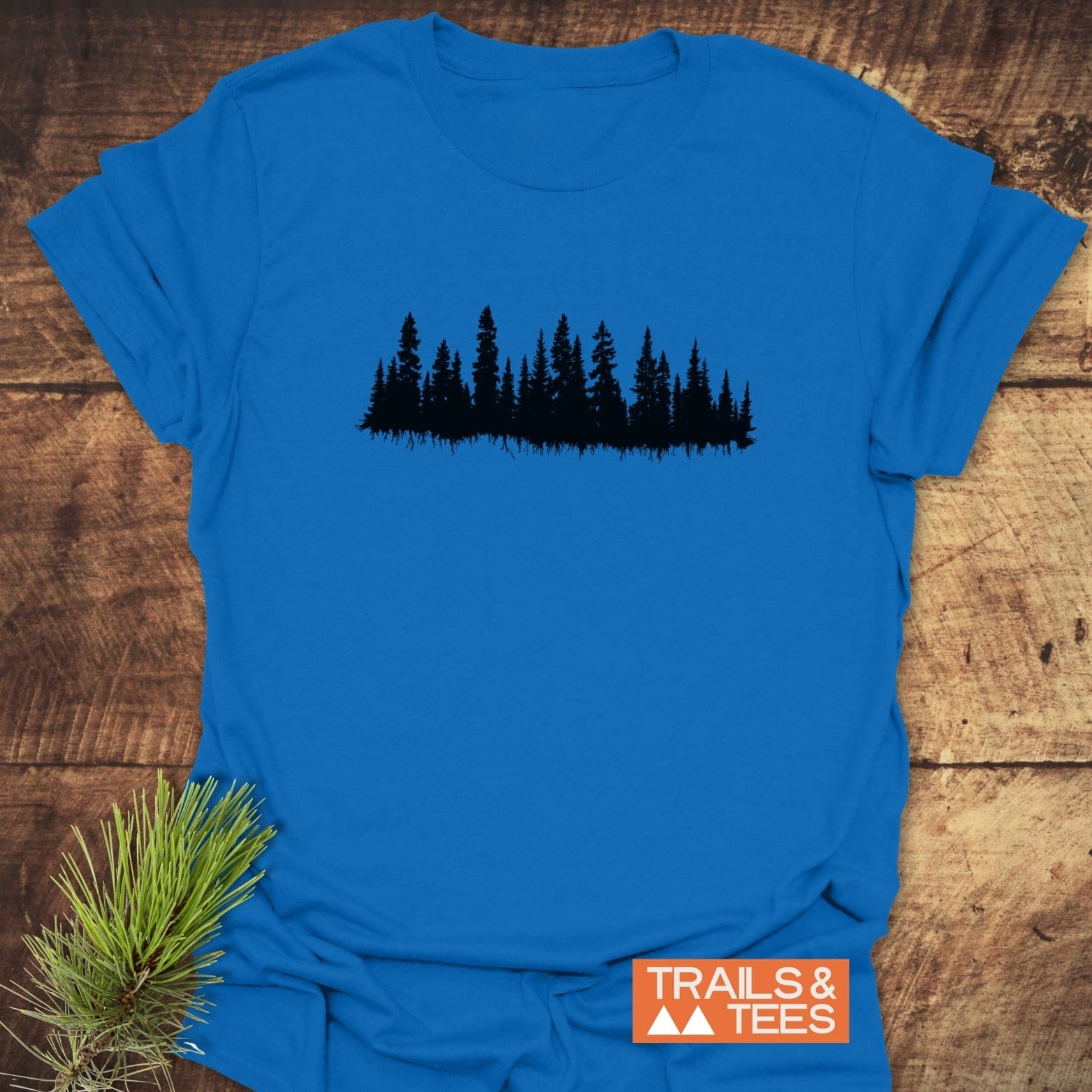 Trees Silhouette T-Shirt with evergreen tree design and Trails & Tees logo, crafted from soft ring-spun cotton for outdoor adventures, laid on wood with pine styling.