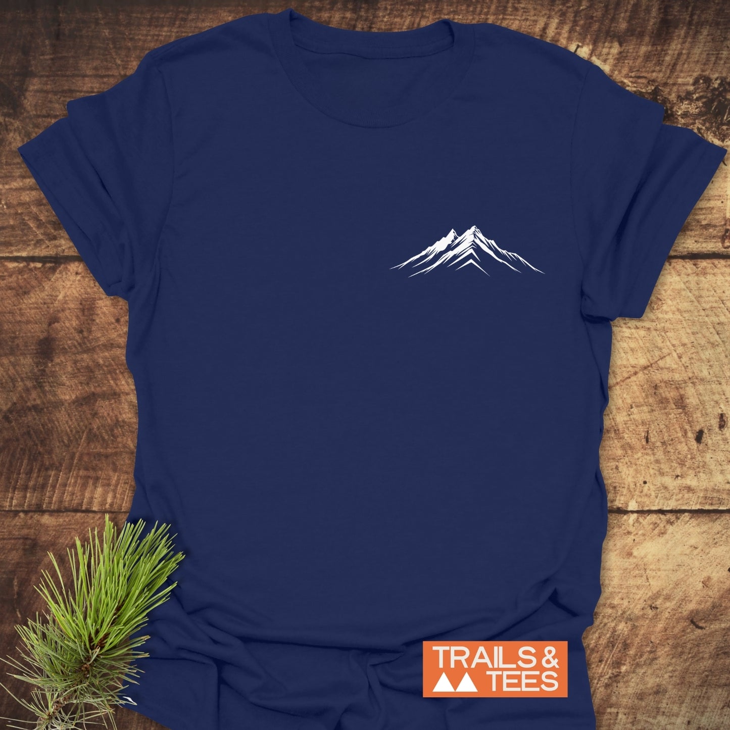 Mountains Logo T-Shirt with a minimalist mountain design on navy-blue fabric, featuring Trails & Tees logo; laid on wood with pine sprig accent.