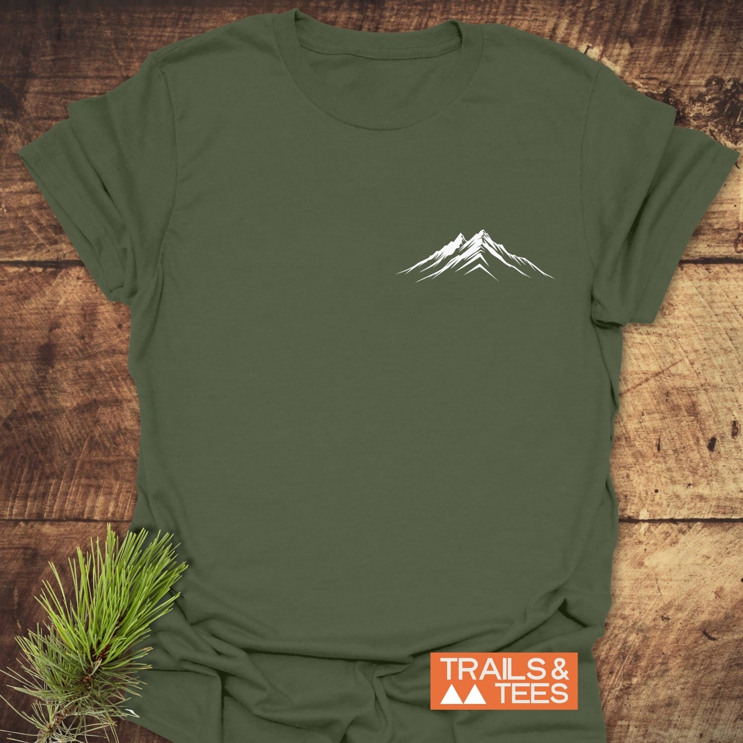 Mountains Logo T-Shirt featuring a white mountain graphic on olive-green fabric, laid flat with TRAILS & TEES logo and pine needles accent.
