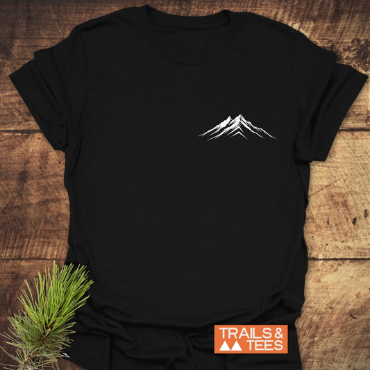 Mountains Logo T-Shirt on wood surface, featuring a white mountain graphic. Includes rustic pine detail and Trails & Tees logo, highlighting outdoor-inspired design.