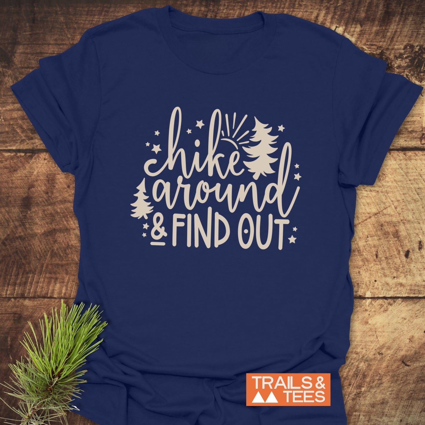 Navy blue Hike Around And Find Out T-shirt with pine trees, stars, and sunburst design, laid on wood, featuring TRAILS & TEES logo and pine sprig.