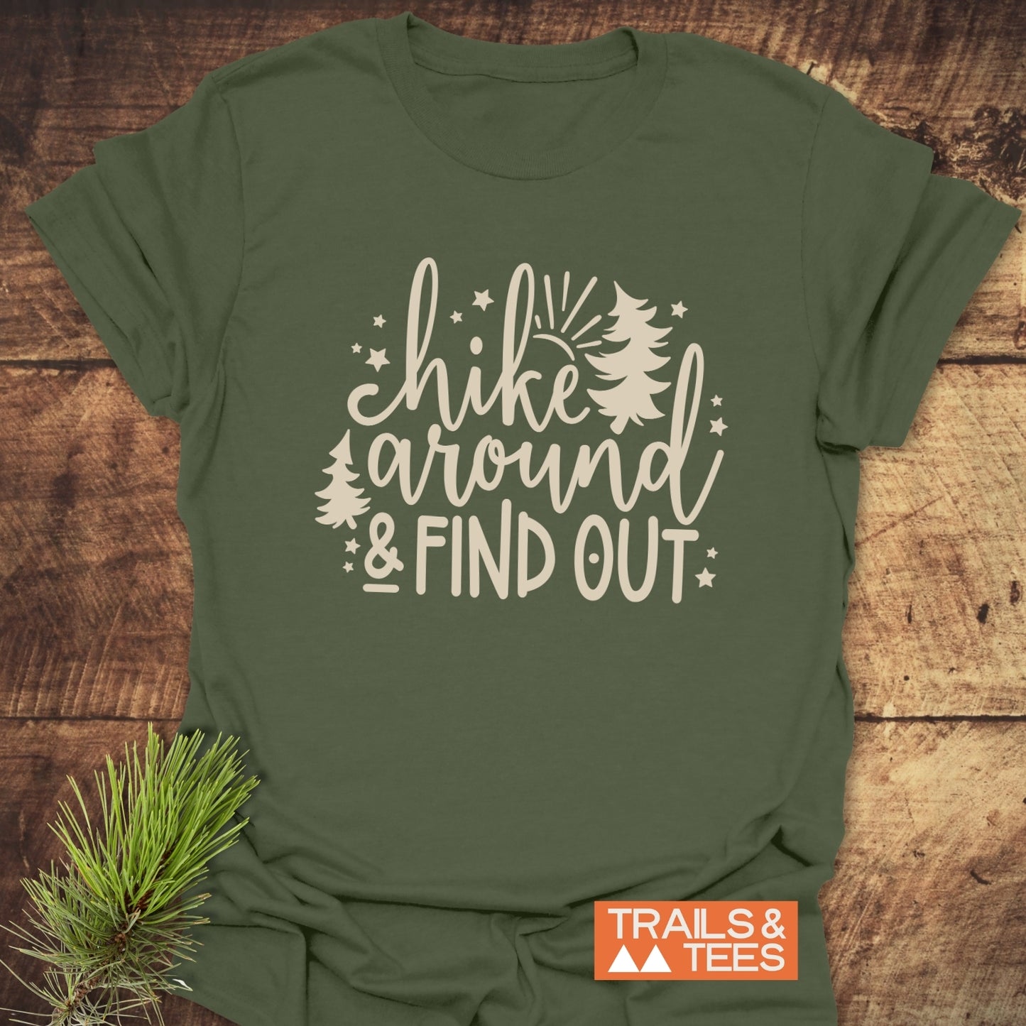 Forest-green T-shirt with Hike Around And Find Out print, featuring pine trees, stars, sunburst, and orange TRAILS & TEES logo on wooden surface.