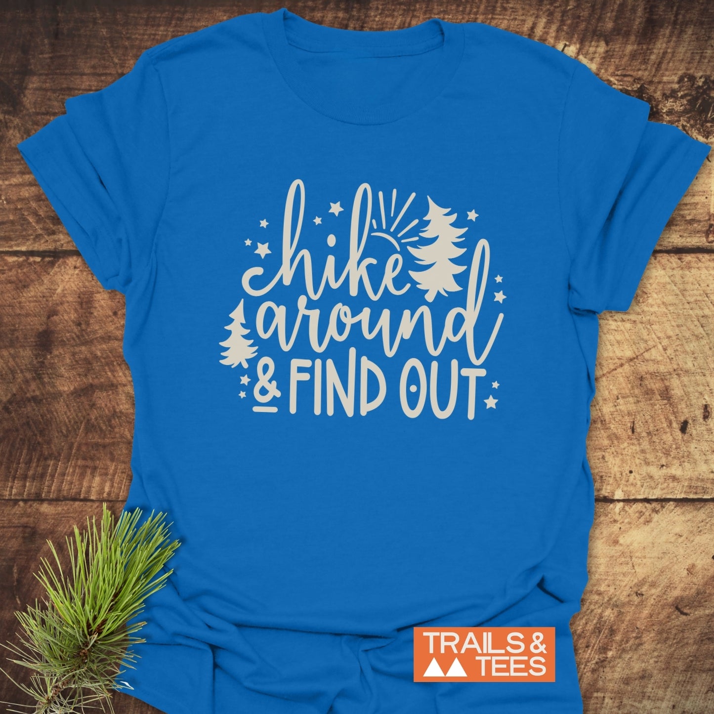 Hike Around And Find Out T-Shirt on rustic wood, featuring white script and nature graphics. Includes Trails & Tees logo; pine sprig accents the scene.