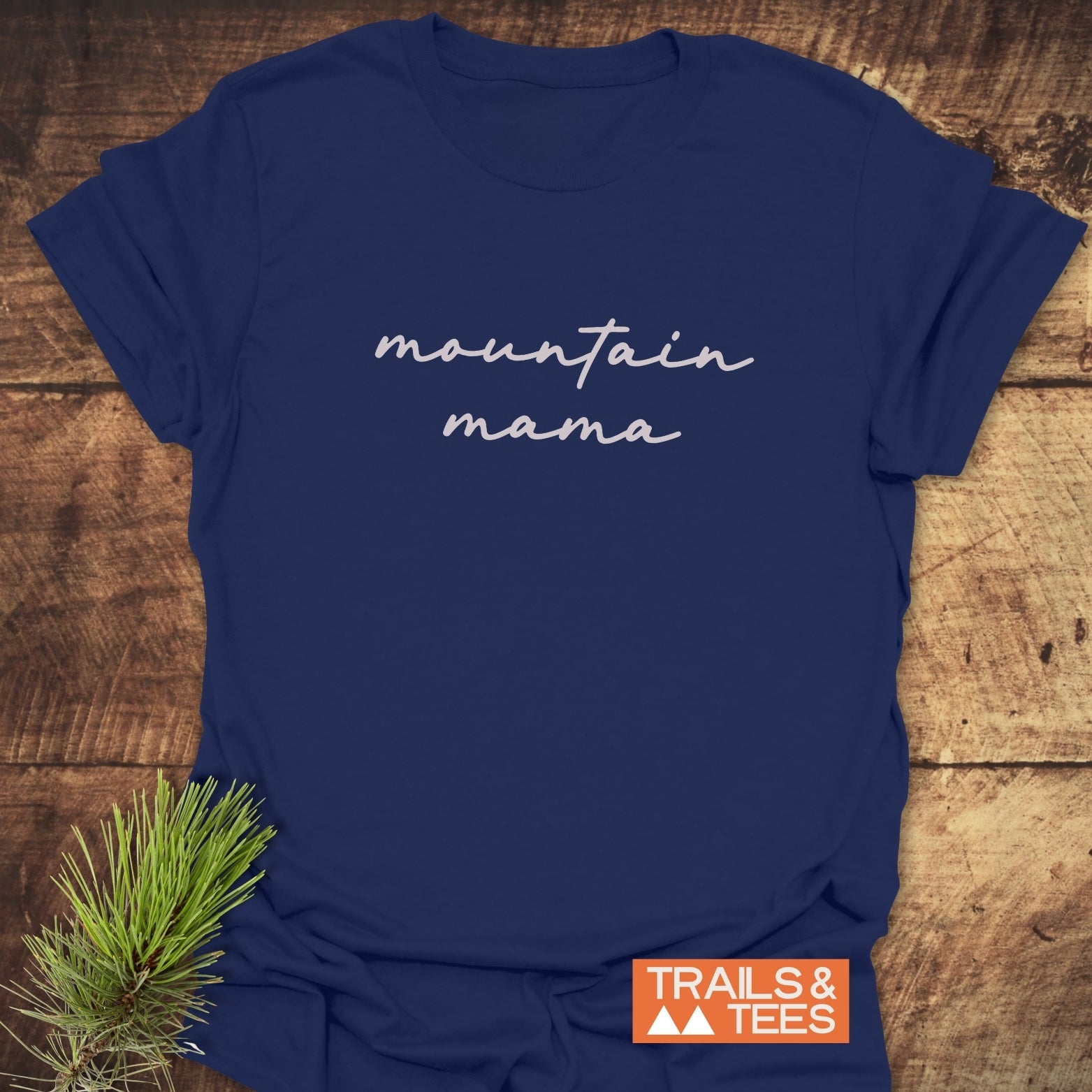 Mountain Mama T-Shirt featuring cursive text on navy-blue fabric, showcasing outdoor spirit. Includes Trails & Tees logo; rustic pine sprig for added charm.
