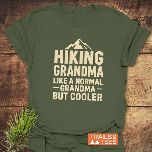 Olive-green Hiking Grandma T-shirt with mountain graphic, crafted from soft cotton for outdoor adventures. Durable print, pre-shrunk, taped shoulders, laid on rustic wood.