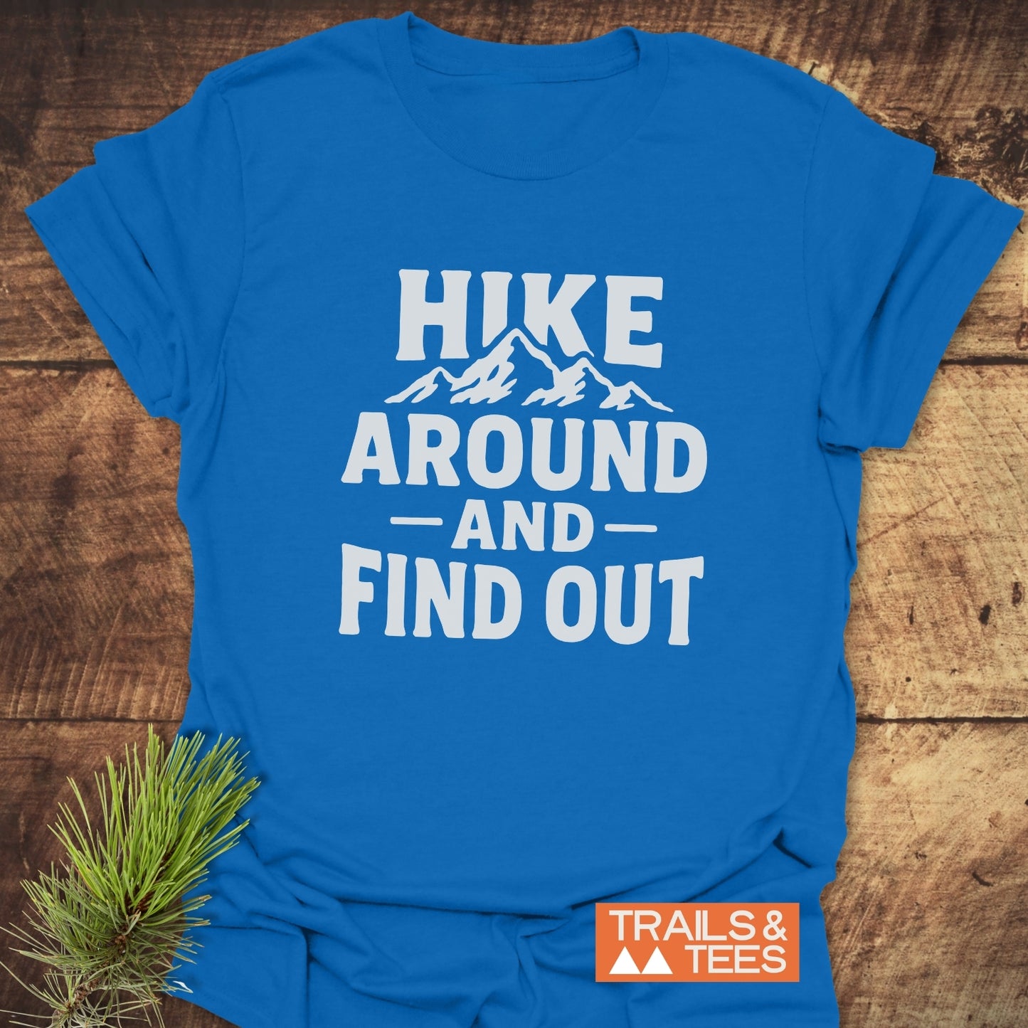 Bright blue Hike Around And Find Out T-shirt with mountain silhouette, laid on wood. Features soft cotton, durable print, designed for outdoor enthusiasts.