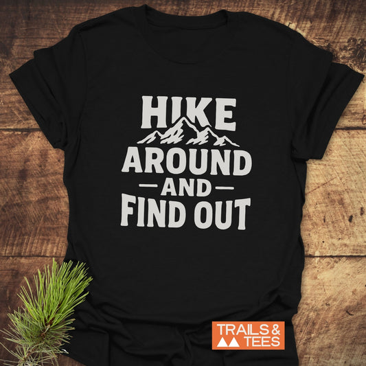 Black Hike Around And Find Out T-Shirt with mountain graphic, showcasing durable, lightweight cotton design ideal for outdoor enthusiasts, laid on wooden surface with pine sprig.