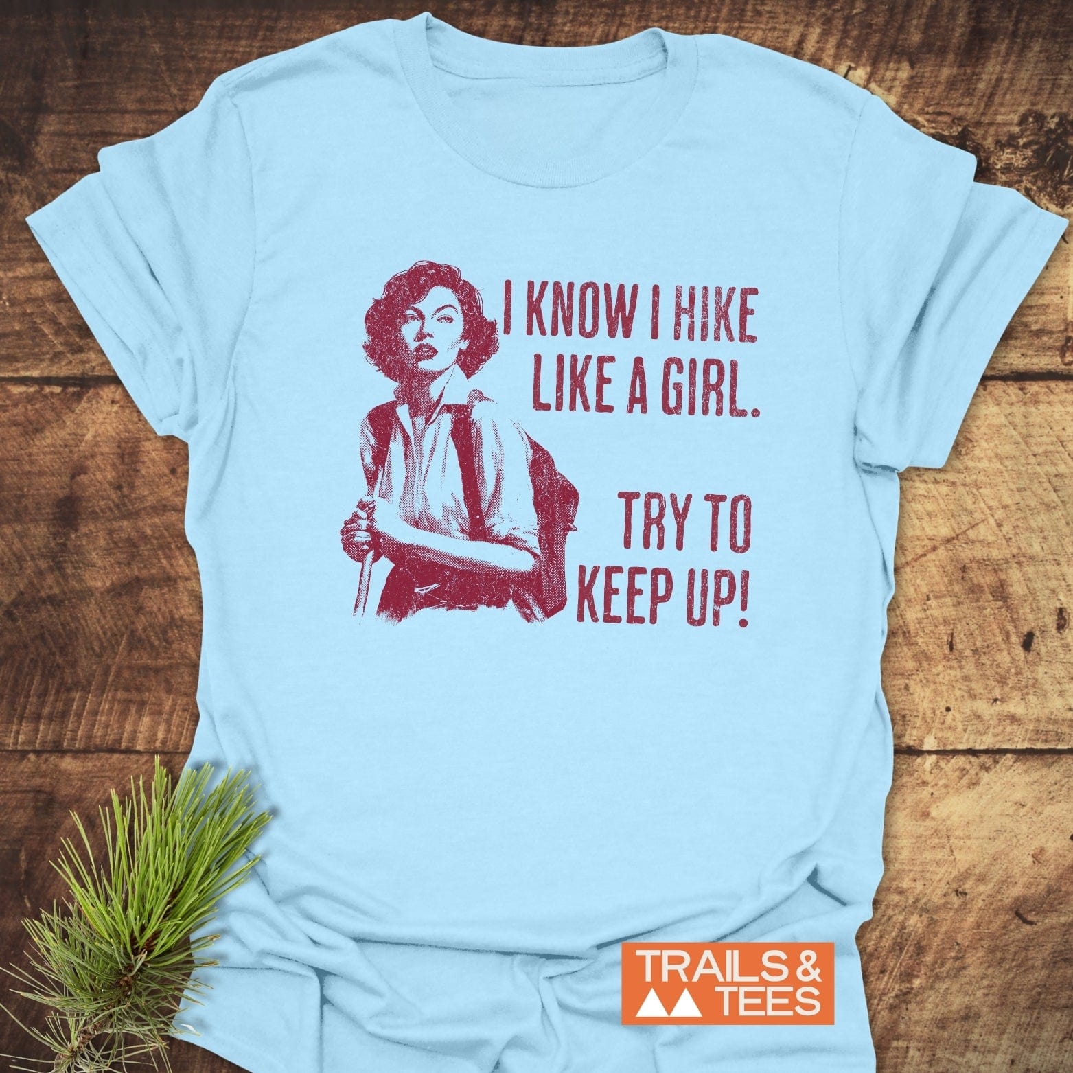 Hike Like A Girl T-Shirt featuring a vintage illustration and slogan, laid on wood with pine sprig; designed for comfort and adventure-focused style.