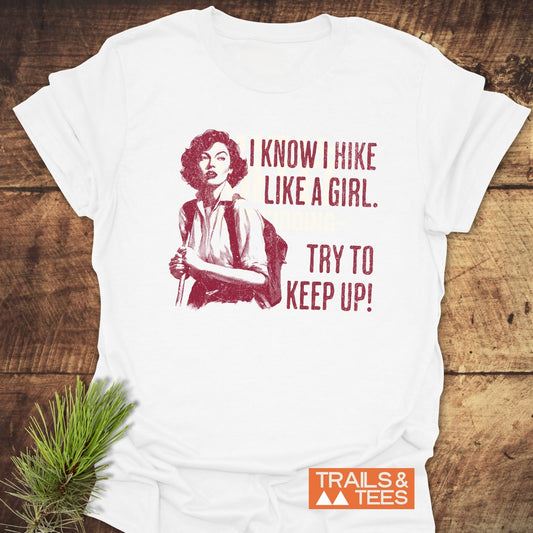 Hike Like A Girl T-Shirt featuring a vintage hiking design, laid on wood with a pine sprig, embodies outdoor adventure spirit and durable comfort.