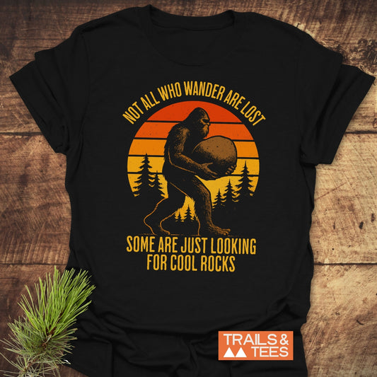 Looking For Cool Rocks T-Shirt features a sunset, pine trees, and Bigfoot graphic with adventure-themed text, crafted from soft, durable cotton for outdoor enthusiasts.