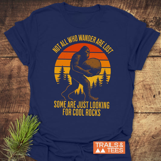Looking For Cool Rocks T-Shirt: Navy-blue tee with Bigfoot silhouette in forest, retro sunset, and adventure quote. Soft cotton, durable print, pre-shrunk for lasting fit.