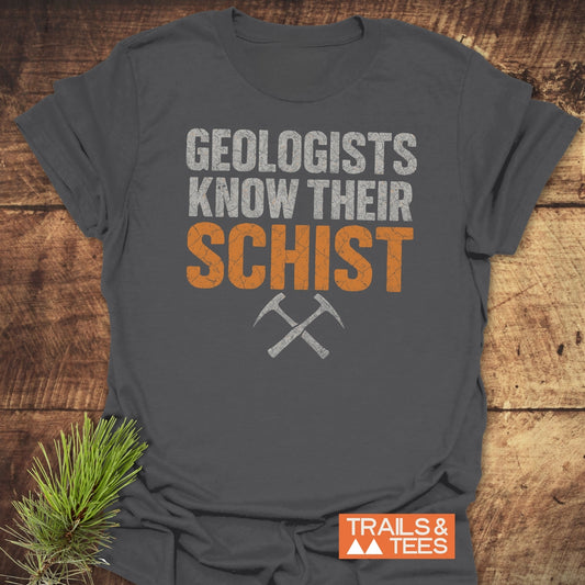 Geologists Know Their Schist T-Shirt featuring a geology pun and rock hammers, laid on wood with pine branch. Made of soft, durable ring-spun cotton.