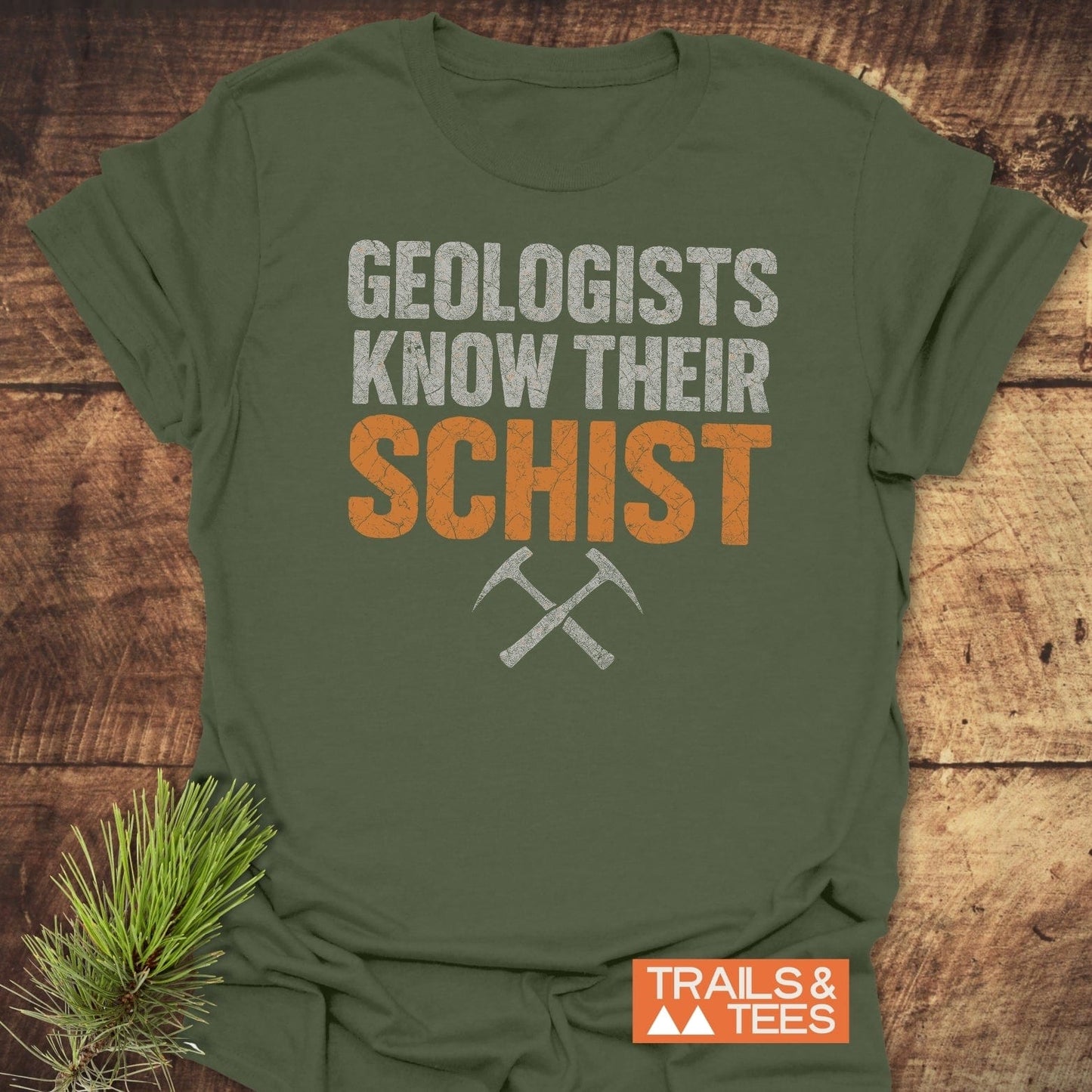 Geologists Know Their Schist T-Shirt with geology pun, crossed rock-hammers, and Trails & Tees logo, laid on wood. Pine sprig adds an outdoor vibe.
