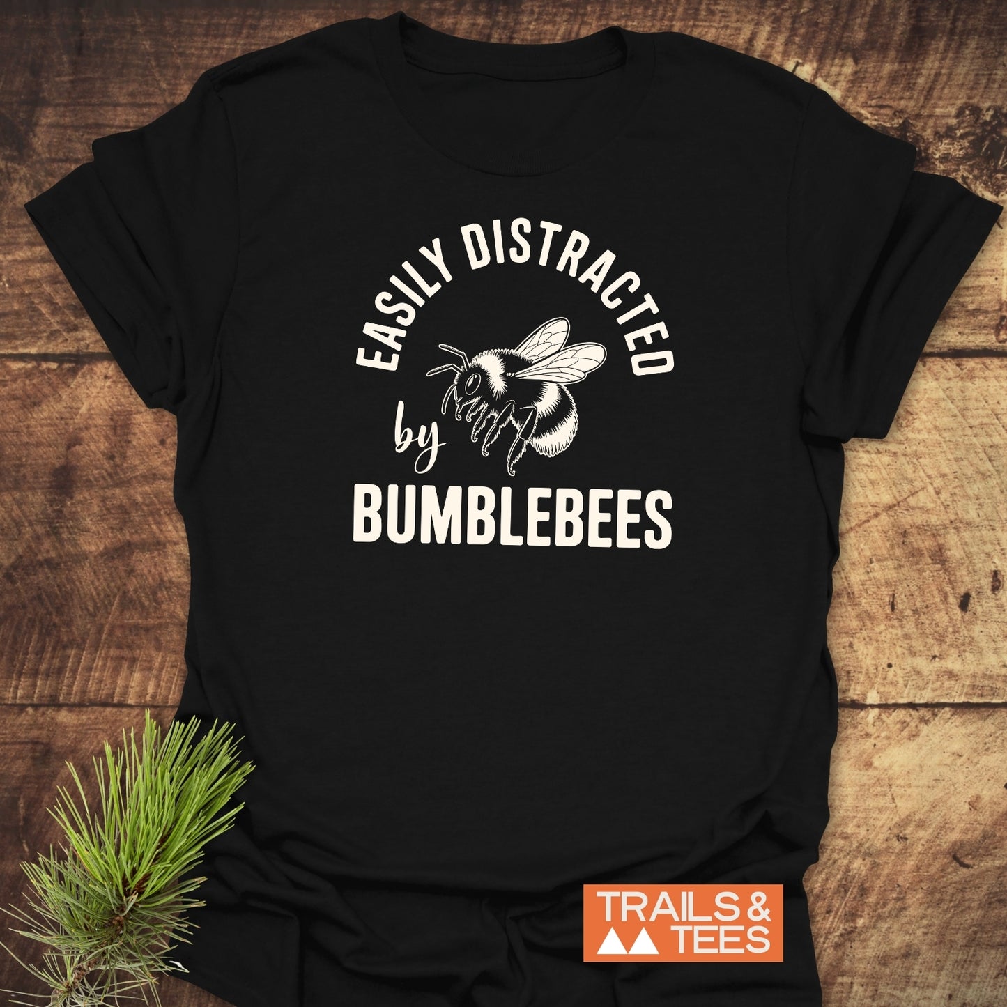 Easily Distracted By Bumblebees T-Shirt on wooden surface, featuring a bumblebee illustration and slogan, styled with pine needles. Made from soft, ring-spun cotton.