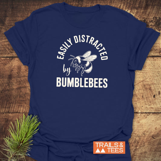 Navy T-shirt with a bumblebee illustration and EASILY DISTRACTED by BUMBLEBEES slogan, styled on rustic wood with a pine sprig, TRAILS & TEES logo visible.