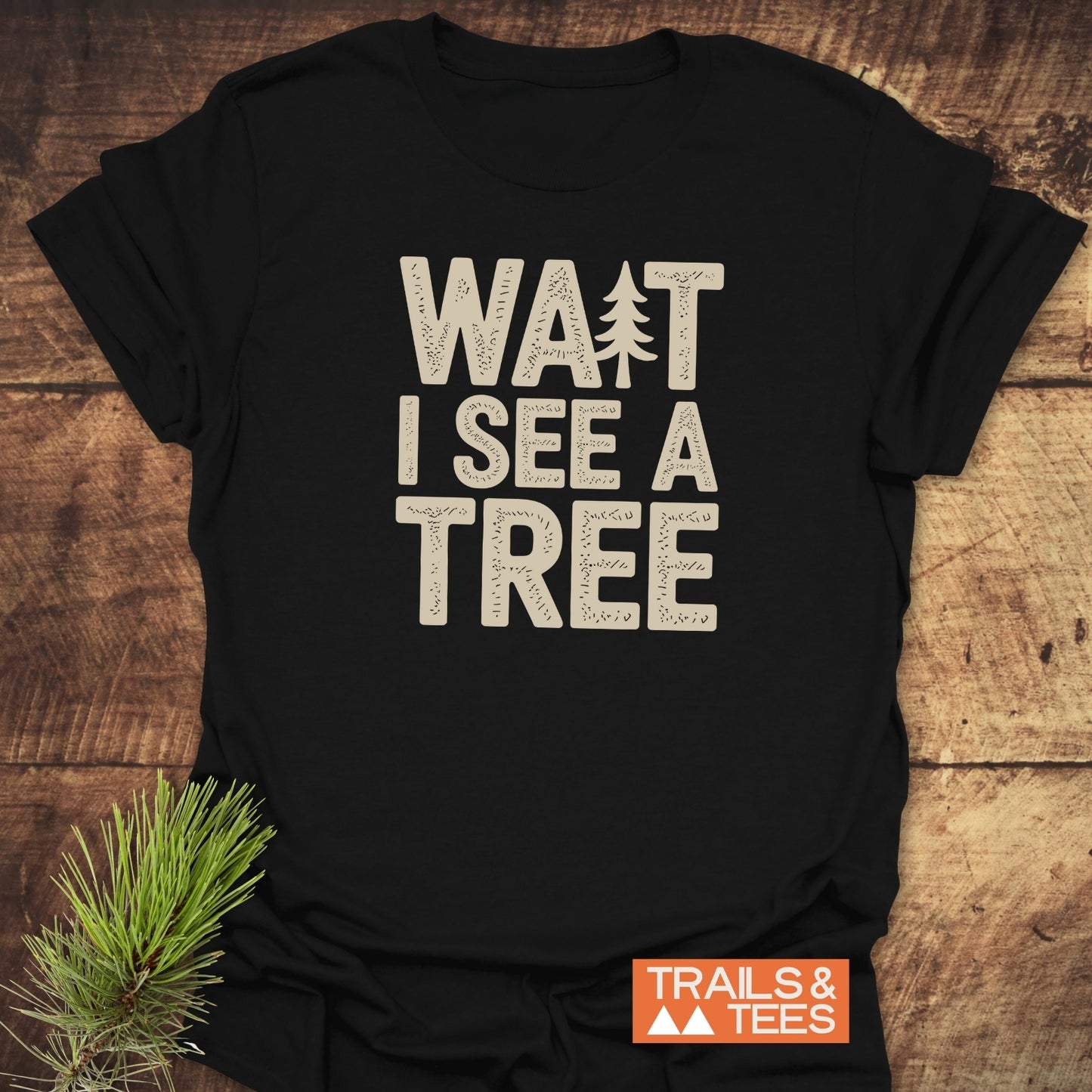 Wait I See A Tree T-Shirt with evergreen icon, laid on wood. Features soft cotton, durable print, and Trails & Tees logo. Includes pine sprig accent.
