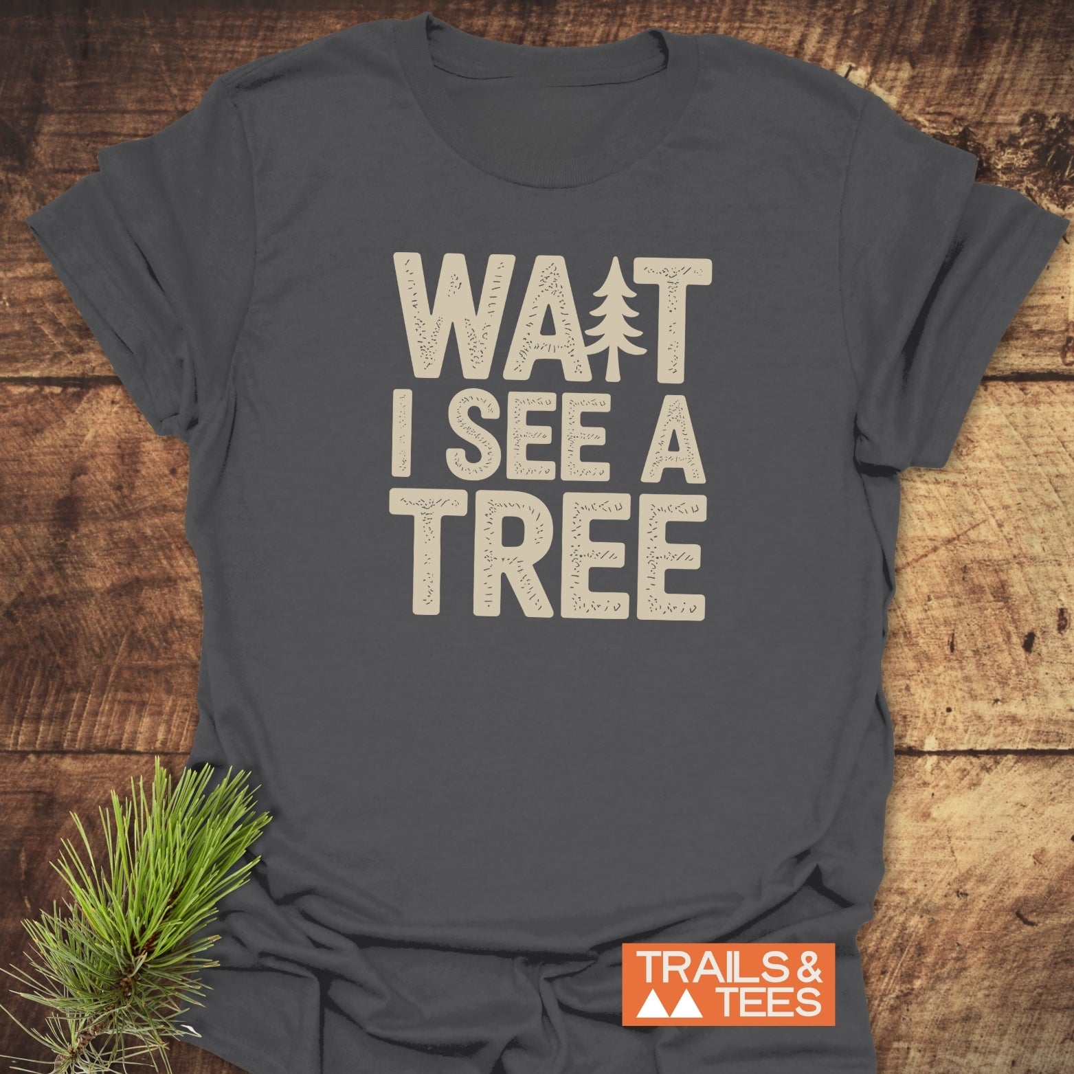 Charcoal-gray Wait I See A Tree T-shirt with cream block letters and tree silhouette, on wood. Includes Trails & Tees logo. Pine sprig decoration.