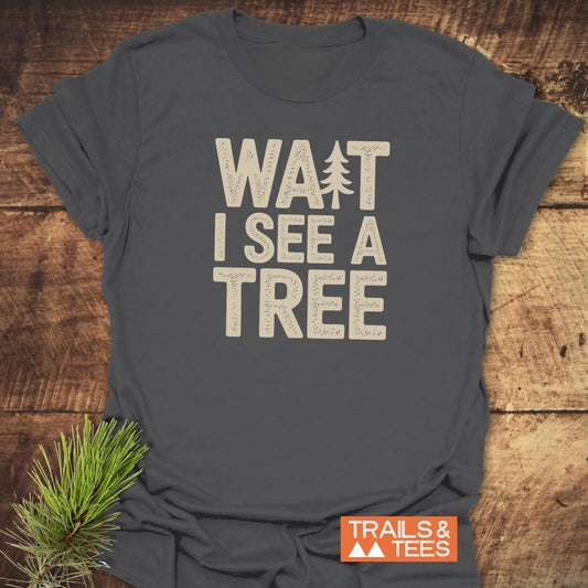 Charcoal-gray Wait I See A Tree T-shirt with cream block letters and tree silhouette, on wood. Includes Trails & Tees logo. Pine sprig decoration.