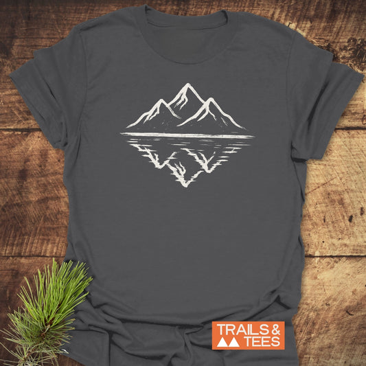 Mountain Reflection T-Shirt featuring white mountain peaks graphic on slate-gray fabric, laid flat with a pine branch and TRAILS & TEES label visible.