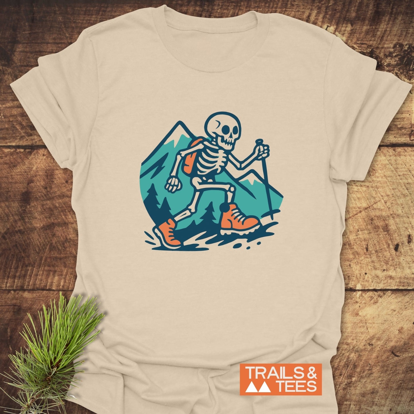 Long Way To The Top – Skeleton Hiking T-Shirt featuring a cartoon skeleton hiking graphic with mountains, emphasizing outdoor adventure and comfort on a flat wooden surface.