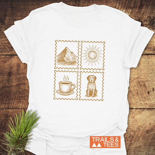 Simple Needs T-Shirt featuring mountain, sun, coffee, and dog illustrations, laid on wood. Made from soft cotton, designed for comfort and outdoor enthusiasts.