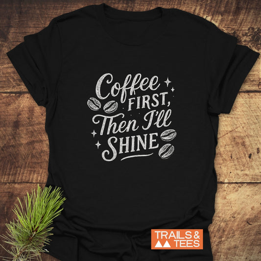 Coffee First T-Shirt with a white hand-lettered design on black cotton, featuring coffee motifs. Laid beside a pine sprig on a wooden surface.