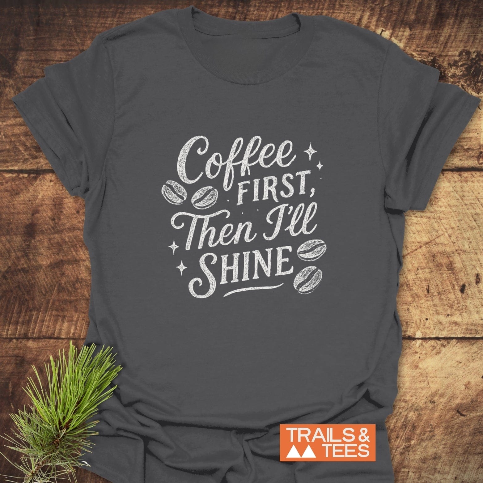 Charcoal-gray Coffee First T-Shirt with white text and coffee bean icons, featuring a small TRAILS & TEES logo, styled with a pine sprig.