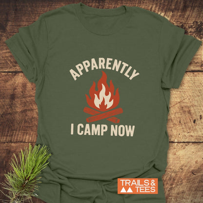 Apparently Camping T-Shirt featuring a campfire graphic and text, displayed on wood. Includes Trails & Tees logo and pine sprig for an outdoorsy theme.