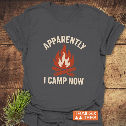 Heather-gray Apparently Camping T-Shirt with campfire graphic and slogan, laid on wood. Features durable print, soft cotton, and rustic pine accent.