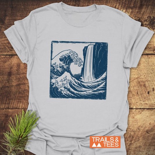 Waterfall And Wave T-Shirt on wood, featuring a navy illustration of a wave and waterfall, styled with a pine sprig. Ideal for outdoor enthusiasts.