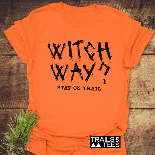 Witch Way? T-Shirt on wood background with pine sprig, featuring durable, brush-stroke print. Perfect for outdoor adventures, crafted from soft, lightweight cotton.