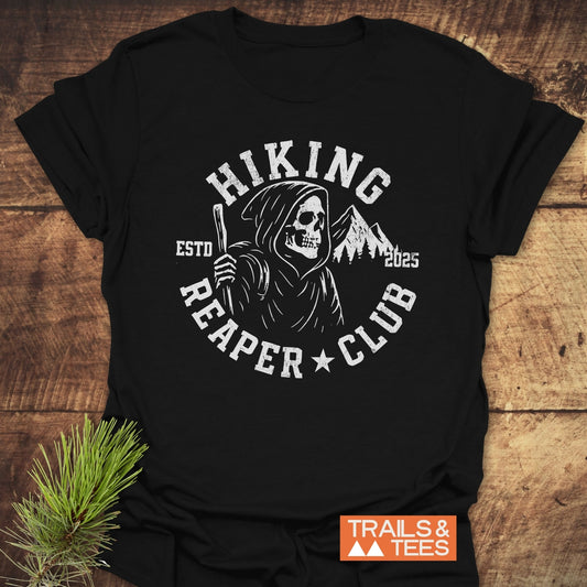 Hiking Reaper Club T-Shirt featuring a white skeletal Reaper with hiking staff and mountains, on black cotton, laid on wood, orange Trails & Tees logo visible.