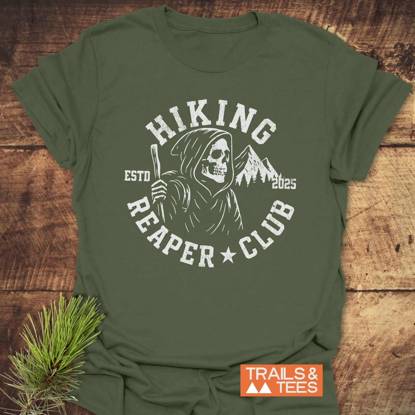 Hiking Reaper Club T-Shirt featuring a Grim Reaper logo with hiking staff and mountains, laid on wood with pine sprig. Durable, lightweight, and pre-shrunk.
