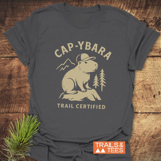 Cap-ybara T-Shirt featuring a capybara graphic with “Trail Certified” text, displayed on a wooden surface with an evergreen sprig for styling.