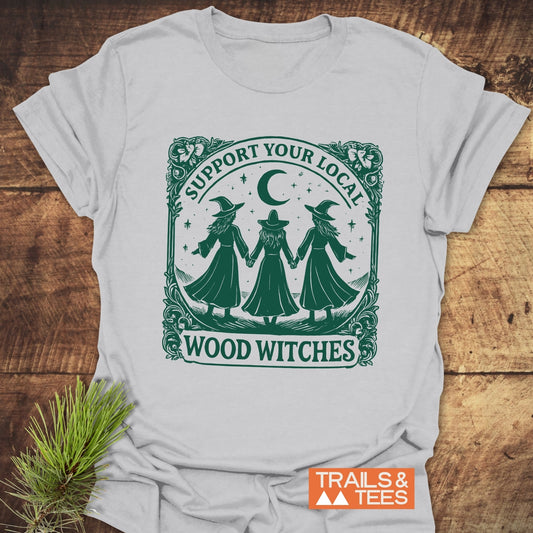 Copy of Take A Hike Human T-Shirt featuring forest-themed witches under crescent moon, with SUPPORT YOUR LOCAL WOOD WITCHES text, laid on wood surface.