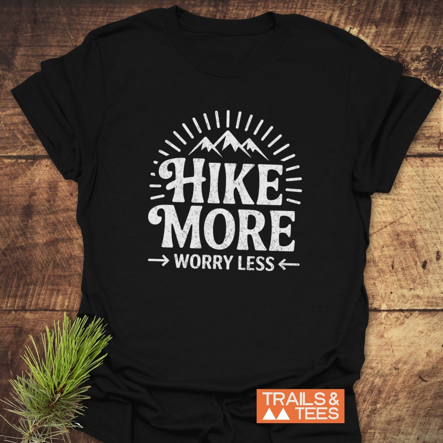 Hike More Worry Less T-Shirt featuring a mountain graphic and slogan, crafted from ring-spun cotton with durable prints, laid on a wooden surface.