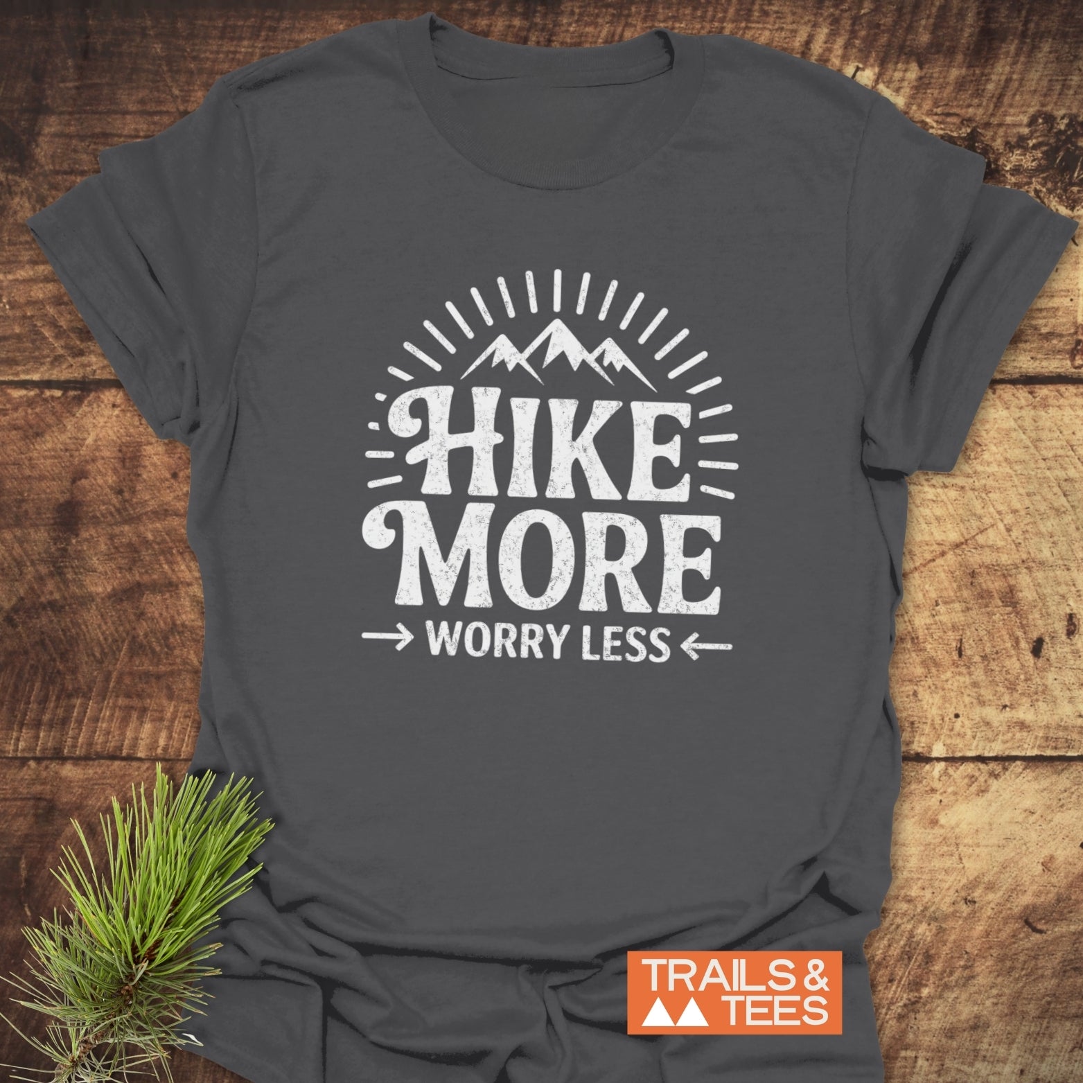 Hike More Worry Less T-Shirt featuring a mountain and sun-ray graphic, laid on wood. Includes “TRAILS & TEES” badge and pine sprig accent.