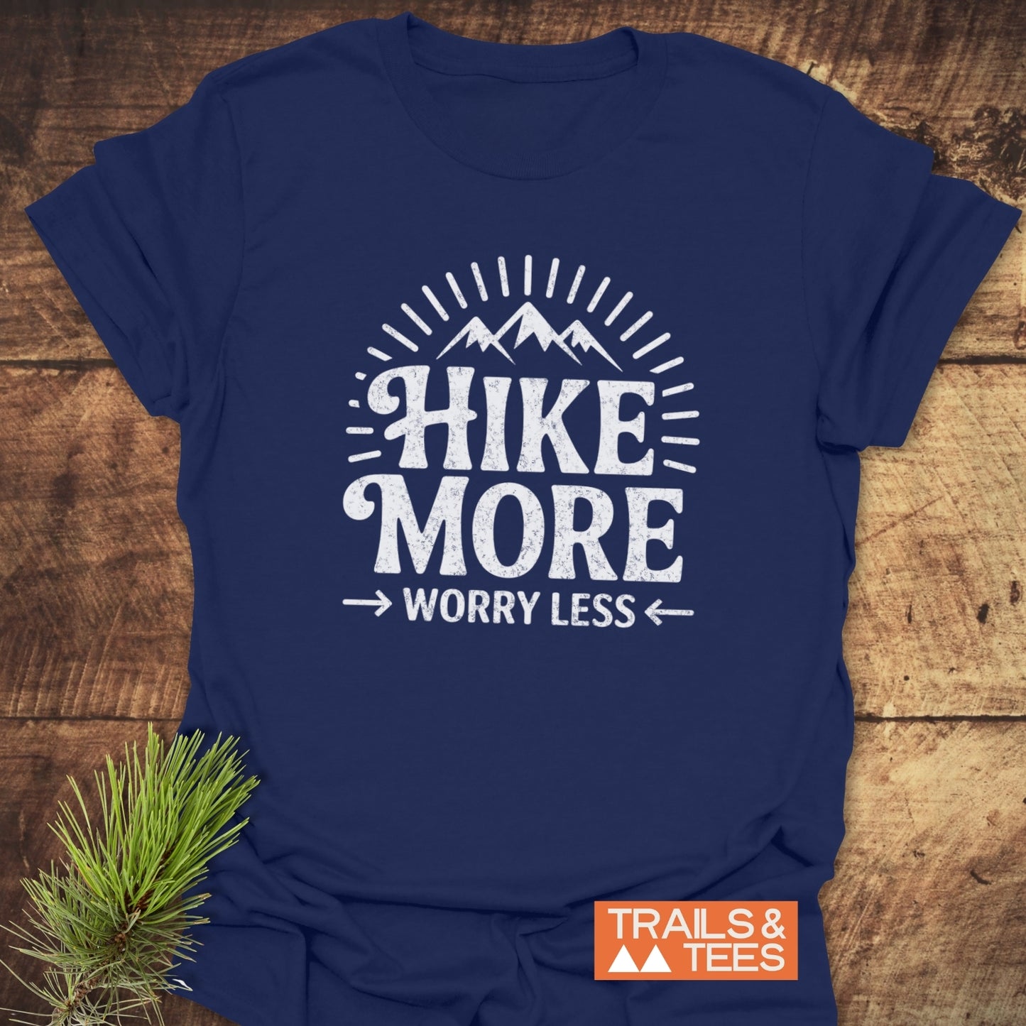 Navy-blue Hike More Worry Less T-shirt with white mountain graphic, laid on wood surface, beside pine sprig, featuring TRAILS & TEES logo in corner.