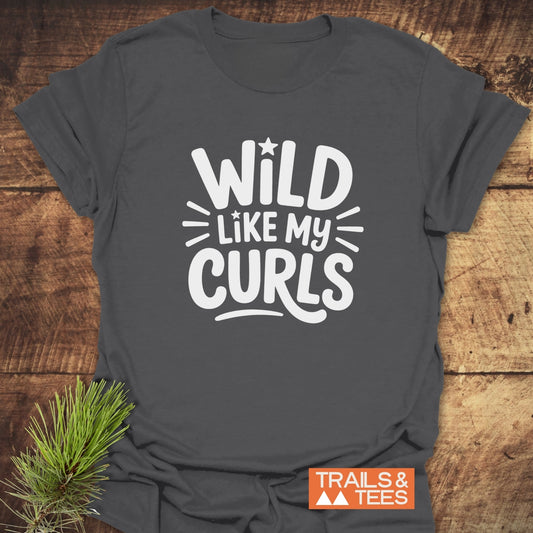 Wild Like My Curls T-Shirt on rustic wood; features white slogan, star accents, orange logo, pine sprig; soft, durable, adventure-ready cotton tee.