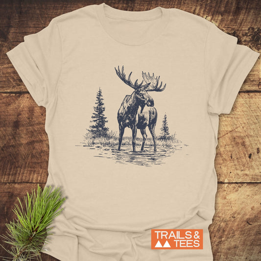 Moose T-Shirt featuring a moose and pine trees in dark-blue, with TRAILS & TEES logo, displayed on wood. Soft, durable, adventure-ready design.