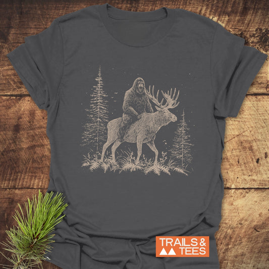 Best Friends Bigfoot T-Shirt featuring Bigfoot riding a moose in a forest, laid on wood with pine sprig. Soft, durable cotton with TRAILS & TEES logo.