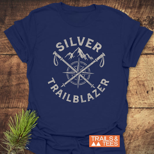 Silver Trailblazer T-Shirt featuring a mountain graphic, compass, and hiking poles, on a wooden surface with pine sprig; durable, soft ring-spun cotton for outdoor enthusiasts.
