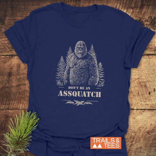 Assquatch T-Shirt featuring Bigfoot art and slogan, laid flat with pine branch; soft, ring-spun cotton, pre-shrunk, durable print, no side seams.