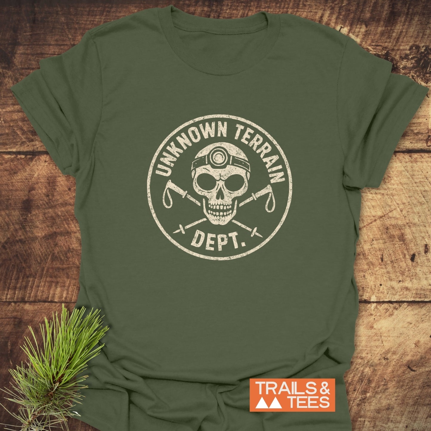 Unknown Terrain Dept. T-Shirt featuring a skull graphic with hiking poles, laid flat with an orange Trails & Tees logo and pine branch accent.