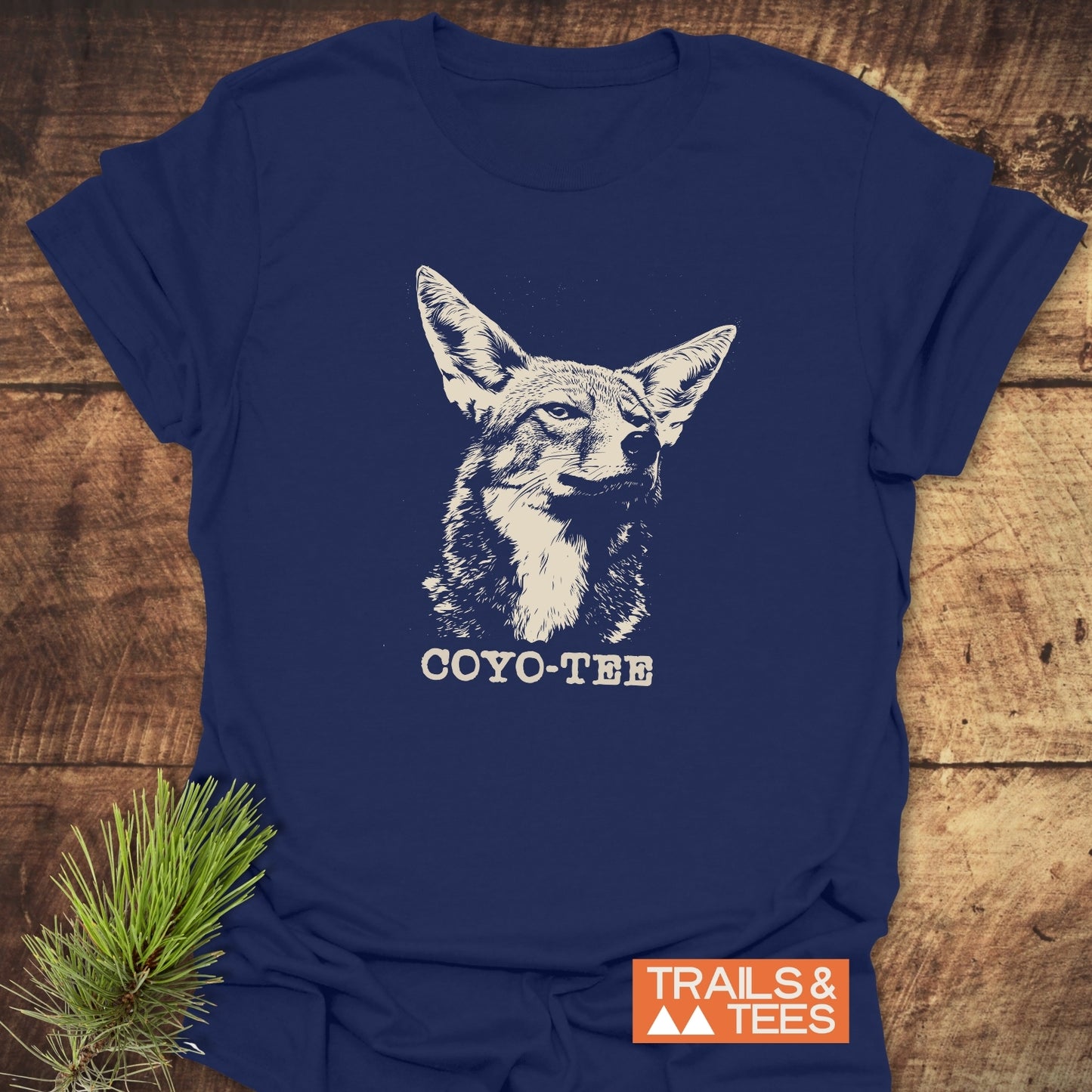 Navy-blue Coyo-Tee T-Shirt with a coyote illustration and “COYO-TEE” caption, featuring a rustic backdrop and Trails & Tees logo on the corner.
